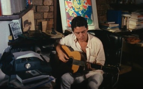 Chico Buarque em um escritório, vestido com roupas claras, toca violão em frente de uma máquina de escrever