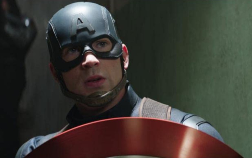 Chris Evans como Capitão América em Capitão América: Guerra Civil, com expressão seria, ele se protege atrás do seu escudo