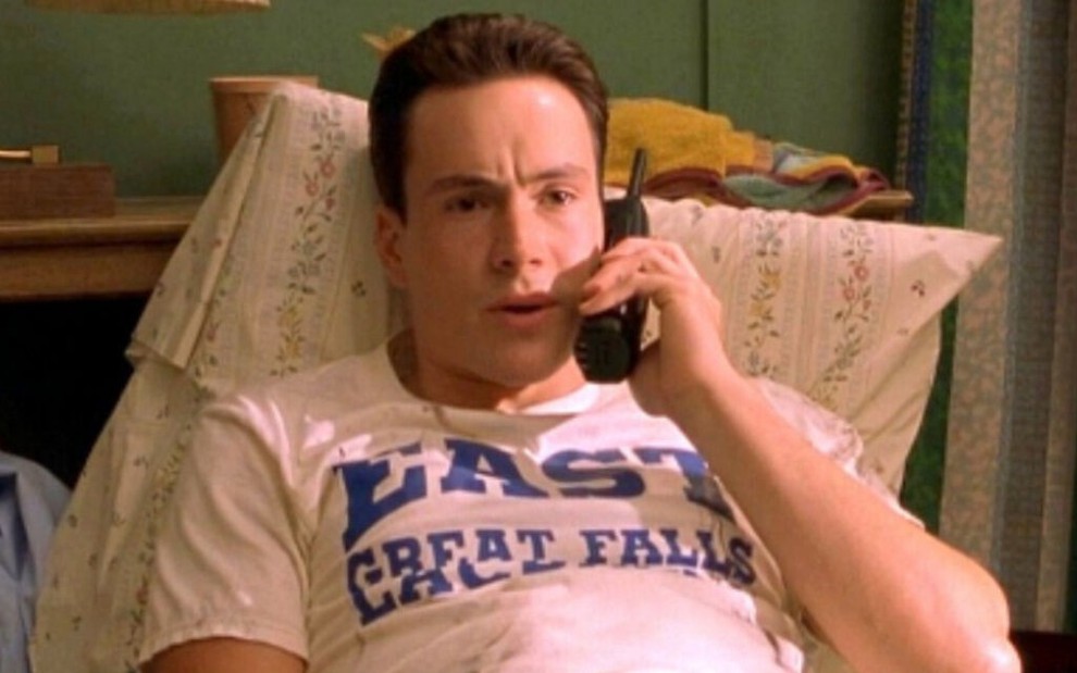 Deitado em uma cama com uma camiseta branca, o ator Chris Klein usa um telefone para conversar sobre sexo em cena do filme American Pie 2