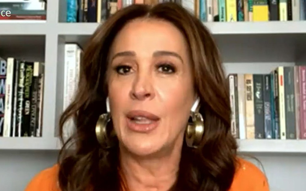 Claudia Raia de blusa laranja, em vídeo chamada de sua casa, com prateleira de livros ao fundo