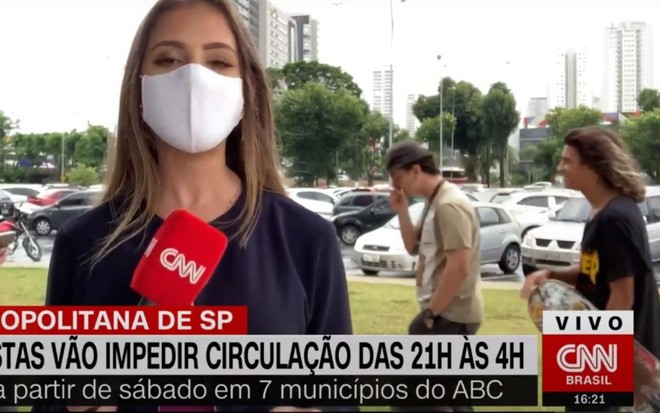 A repórter Manuella Niclewicz durante reportagem; dois jovens passam ao fundo, e o primeiro leva a mão à boca para tossir