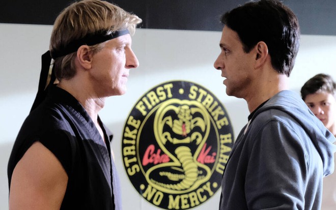 Dentro de um dojo, o loiro William Zabak, com uma faixa preta na cabeça, encara o moreno Ralph Macchio em Karatê Kid