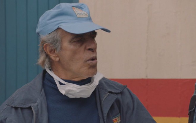 O ator Walter Breda, com boné azul e uma máscara branca no queixo, faz biquinho em cena da comédia nacional Auto Posto