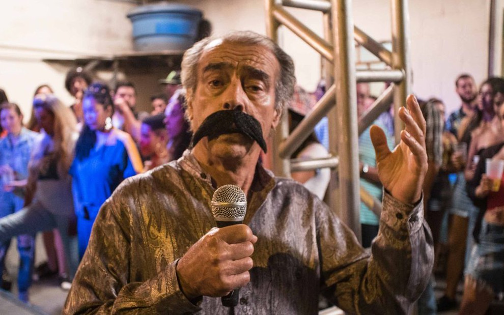 Com um bigode falso, camisa estampada e microfone na mão, Walter Breda posa como cantor brega em cena de Auto Posto