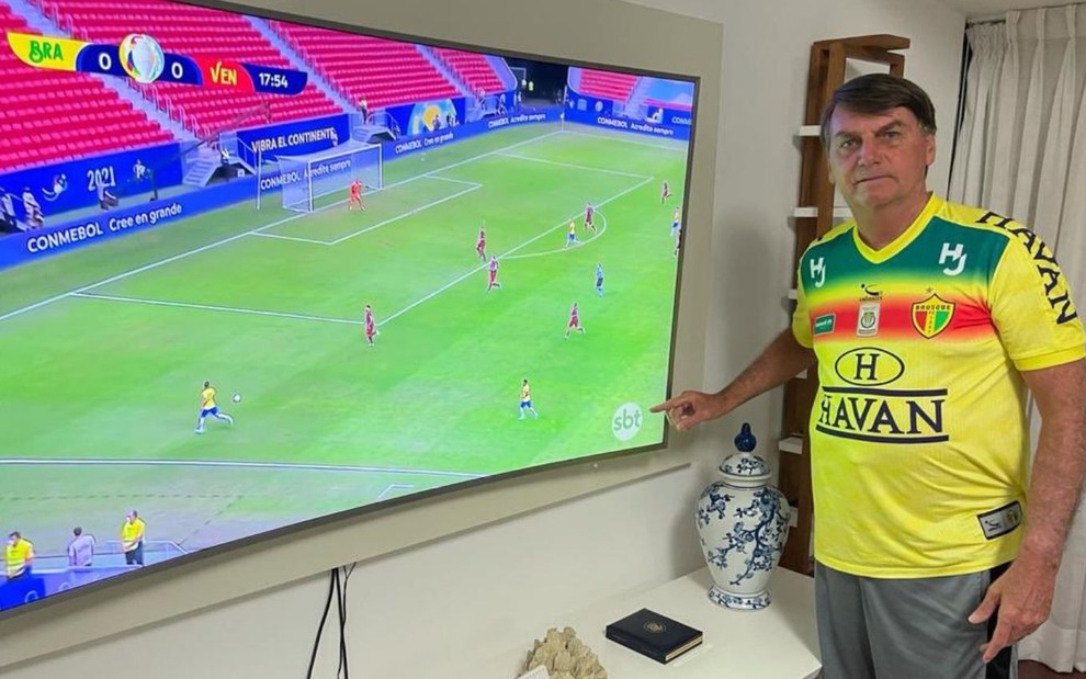 Com camiseta de time apoiado pela Havan, Jair Bolsonaro aponta para o logo do SBT em jogo de estreia da Copa América