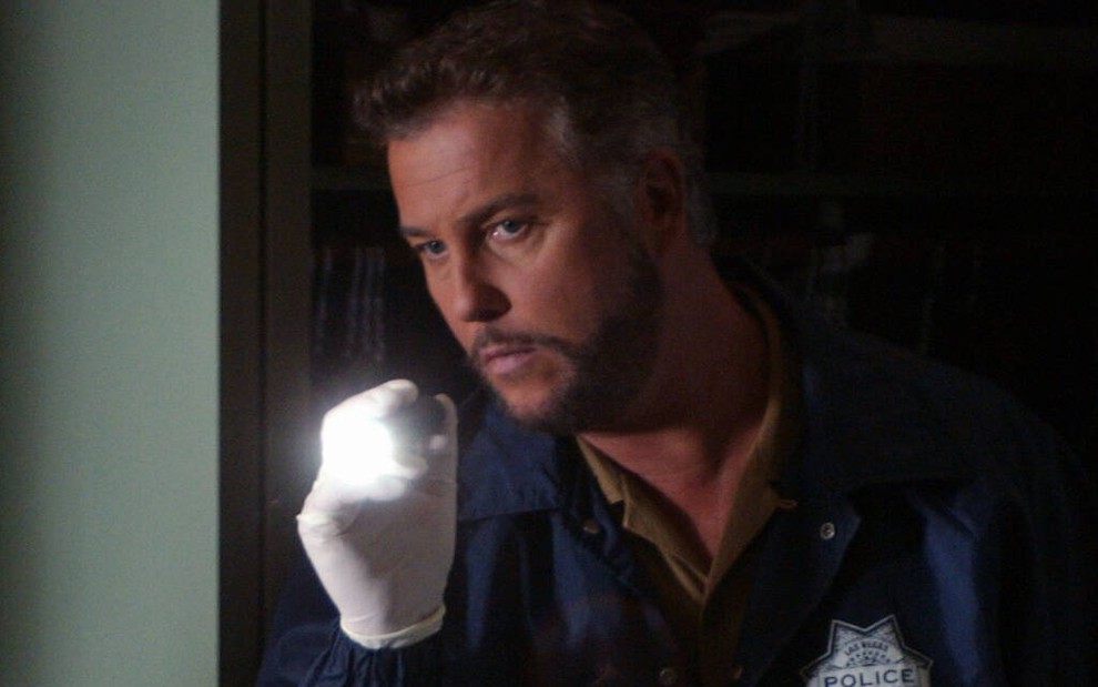 Em um ambiente escuro, William Petersen usa uma luva branca e acende uma lanterna em cena clássica de CSI