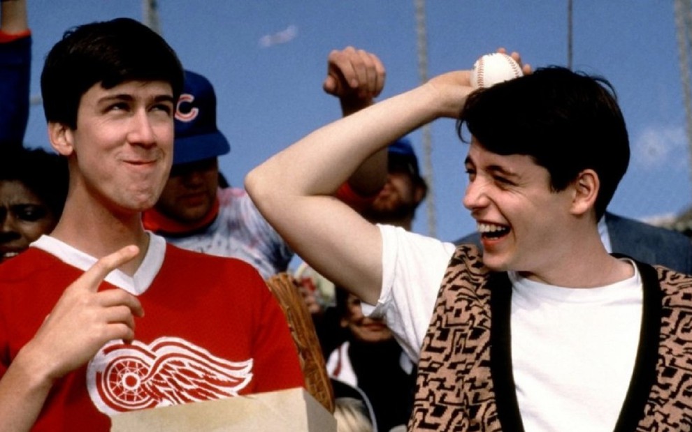 Cameron (Alan Ruck), de camiseta vermelha, e Ferris (Matthew Broderick), de camiseta branca e colete, sorriem na arquibancada enquanto assistem a um jogo de beisebol
