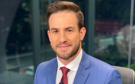 Daniel Adjuto sorri discretamente e usa terno em foto que aparece nos estúdios da CNN Brasil