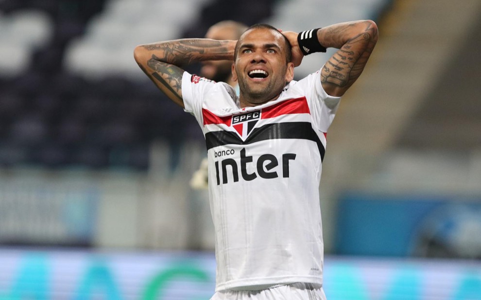Daniel Alves em campo pelo São Paulo, vestido com uniforme do time nas cores branco, preto e vermelho, o jogador está com as mãos atrás da cabeça e expressão de indignação.
