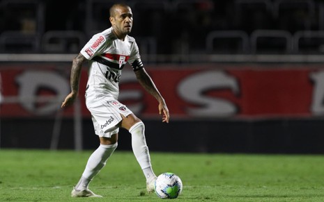 O jogador Daniel Alves vestido com o uniforme branco do São Paulo, tocando um bola de futebol com o pé