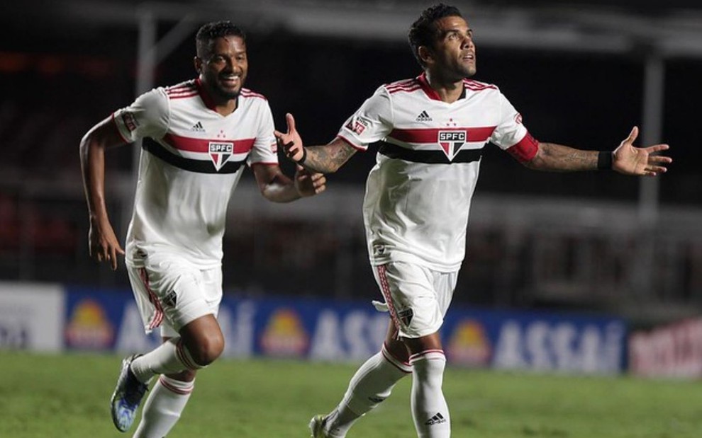 O jogadores do São Paulo, Reinaldo e Daniel Alves, comeram gol do time em campo, correm de braços abertos, vestidos com o uniforme do time nas cores branco, vermelho e preto.
