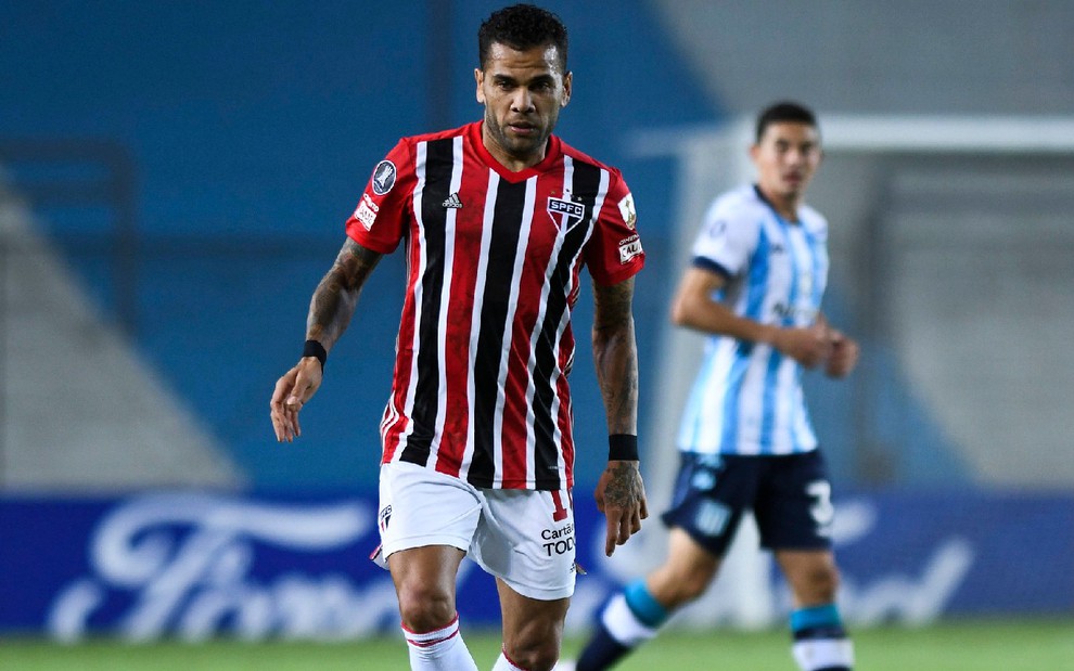 Daniel Alvez com a camisa vermelha, preta e branco do São Paulo observado por adversário desfocado ao fundo