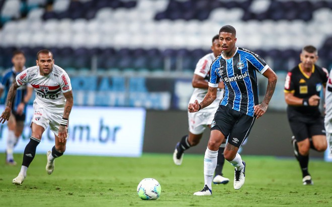 Daniel Alves, do São Paulo, e Jean Pyerre, do Grêmio, em disputa de bola no campo; Daniel Alves veste o uniforme do São Paulo nas cores branco, preto e vermelho, Jean Pyerre veste o uniforme do  Grêmio nas cores azul, preto e branco
