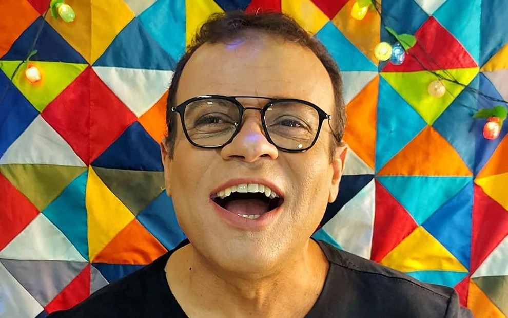 Daniel está em frente a uma parede colorida (azul, vermelho, verde, laranja e branco), ele está de boca aberta, camiseta preta e óculos de grau