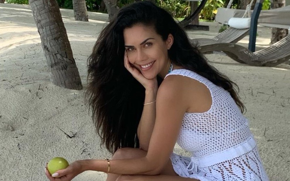 Daniela Albuquerque com os cabelos soltos em uma praia, com fruta na mão e vestido branco