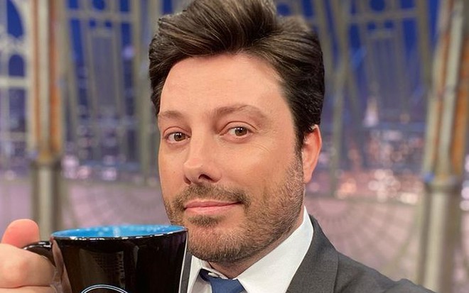 Danilo Gentili de terno preto, gravata azul, segurando a caneca do The Noite