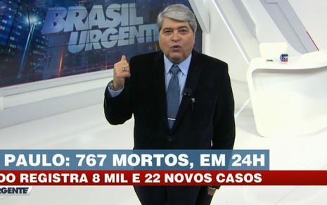 José Luiz Datena com um terno escuro, camisa e gravata azuis, com a mão direita erguida em um cenário de programa de TV