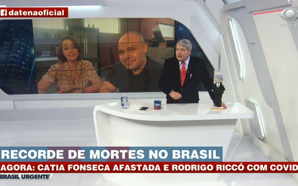 José Luiz Datena ao lado de miniatura sua em boneca de pano com Catia Fonseca e Rodrigo Riccó em telão atrás dele no Brasil Urgente