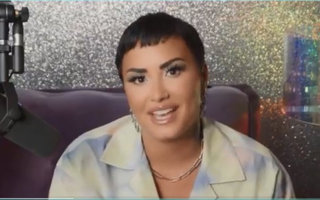 Demi Lovato com blazer claro e cabelo curto olhando para a câmera sentada em frente a parede prateada cintilante