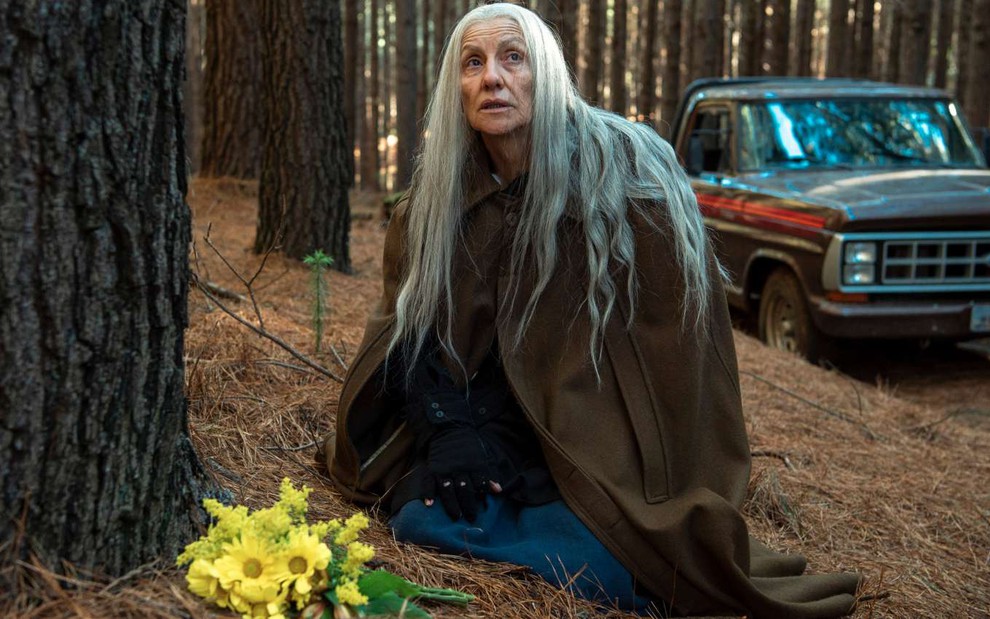 A atriz Cassia Kis com longos cabelos brancos, em floresta, em frente a buquê de flores amarelas, olha para cima em cena de Desalma