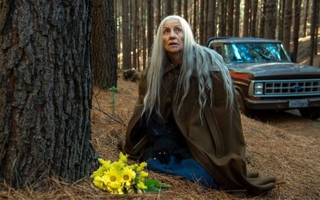 Cássia Kis caracterizada como uma feiticeira em Desalma; atriz está abaixada na floresta com flores amarelas a sua frente