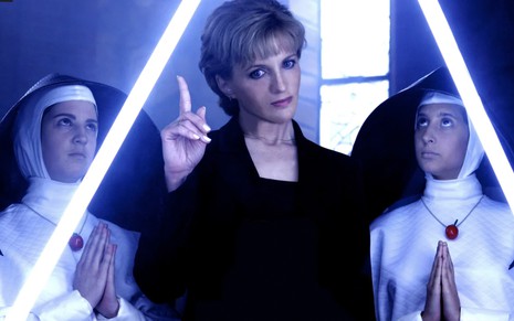 Entre duas freiras, uma sósia de Diana chamada Denise Watson ergue o dedo indicador da mão direito e aponta para o teto em cena de Diana: The Rose Conspiracy