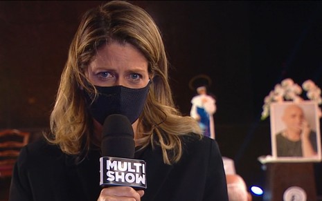 Didi Wagner chora enquanto olha para câmera, com microfone do canal Multishow; ao fundo, um quadro de Paulo Gustavo desfocado