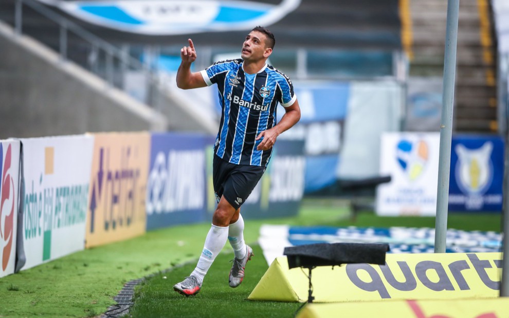 O centroavante Diego Souza comemora gol pelo Grêmio, vestido com o uniforme do time nas cores azul e preta, o jogador corre atrás do gol com o dedo indicador de uma das mãos levantado