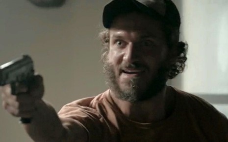 O ator Paulo Rocha, com expressão de descontrolado, aponta uma arma de fogo para frente em cena como Dino de Totalmente Demais