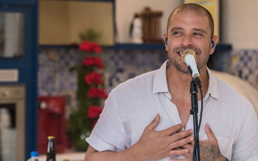 Diogo Nogueira sorrindo de camisa branca com as mãos no peito em frente a um microfone