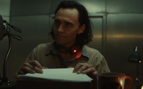 Tom Hiddleston está com uma coleira eletrônica e segura uma caneta e um formulário em cena de Loki