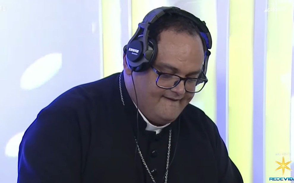 Padre aparece em frente a um fundo branco e está com um fone de DJ, óculos, enquanto toca a picape