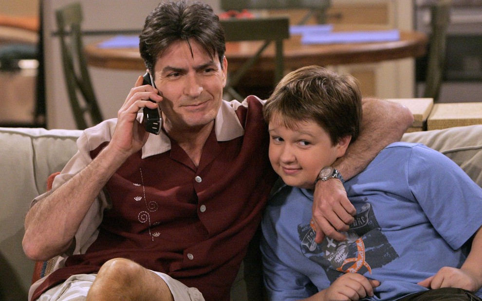Com um telefone na orelha, Charlie Sheen abraça Angus T. Jones pelo pescoço em cena de Dois Homens e Meio