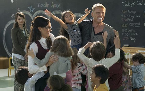 Os atores Dolph Lundgren, Sarah Strange e Darla Taylor rodeados de crianças em uma sala de aula no filme Um Tira no Jardim de Infância 2