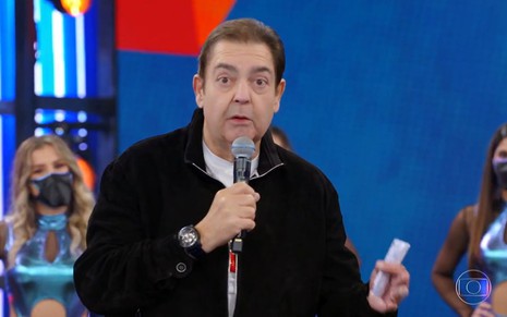 Fausto Silva de jaqueta preta e microfone na mão na apresentação do Domingão do Faustão; uma bailarina de máscara aparece ao fundo