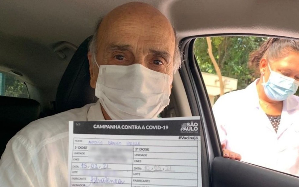 Drauzio Varella de máscara segurando o cartão de vacinação de dentro do carro