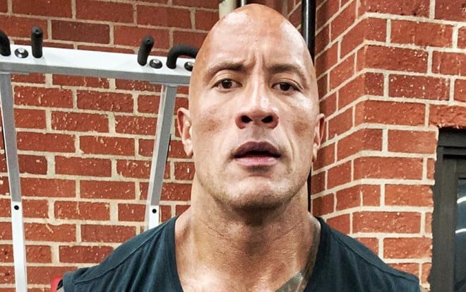 Dwayne Johnson suado e de boca aberta em um cenário com aparelho de ginástica ao fundo