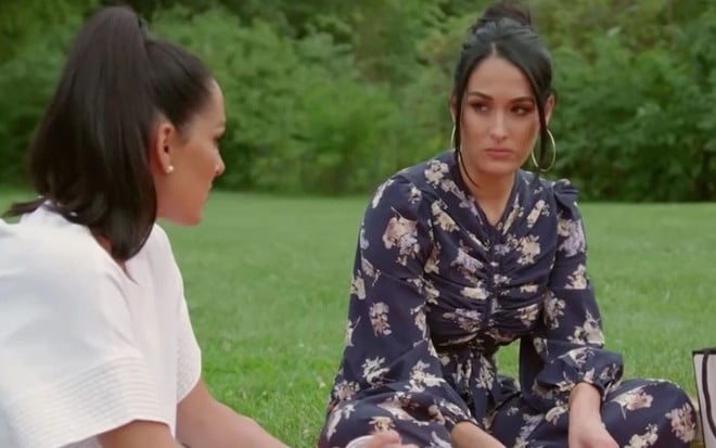 As gêmeas Brie Bella (de branco) e Nikki Bella (de azul com estampa) conversam sentadas em um gramado, estilo piquenique