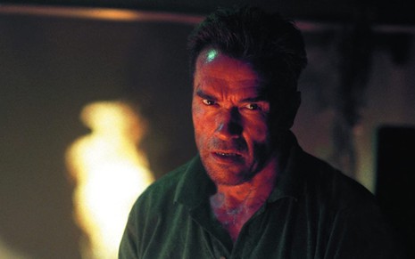 Arnold Schwarzenegger olhando para frente com uma feição séria, suando, enquanto alguma coisa pega fogo em segundo plano