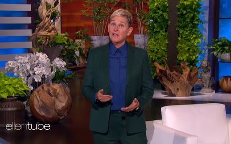Elle DeGeneres veste terno verde escuro e camisa azul no comando de seu programa no palco com diversos detalhes de decoração no fundo e olha para frente