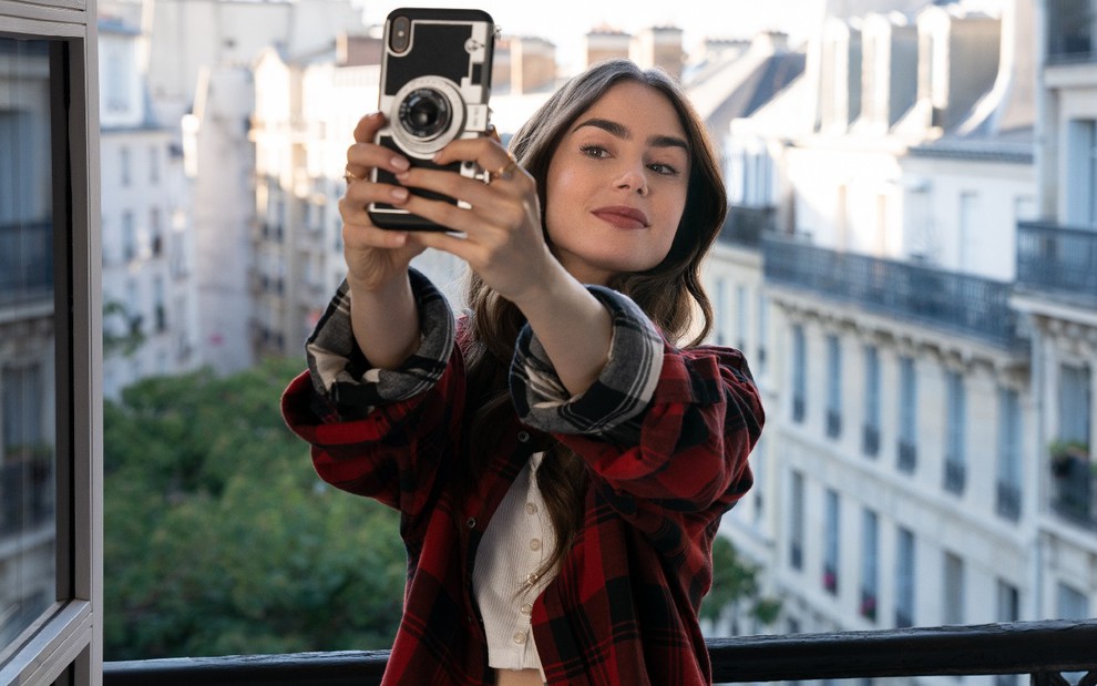 Lilly Collins de camisa branca e blusa xadrez vermelha segurando o celular com as duas mãos esticadas para fazer foto de si mesma em uma janela aberta com prédios aos fundo