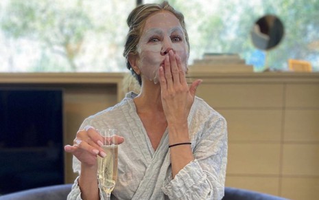 Imagem de Jennifer Aniston de roupão, segurando uma taça de champanhe e usando máscara facial
