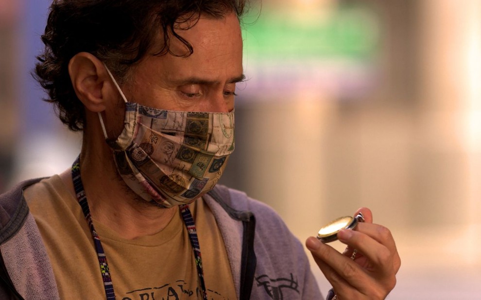 Enrique Diaz, caracterizado como Durval, de Amor de Mãe, olha para relógio usando máscara