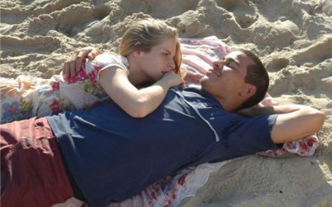Thiago Martins deitado em cima de uma canga na areia, e Vitória com o rosto e o braço apoiado em seu peito