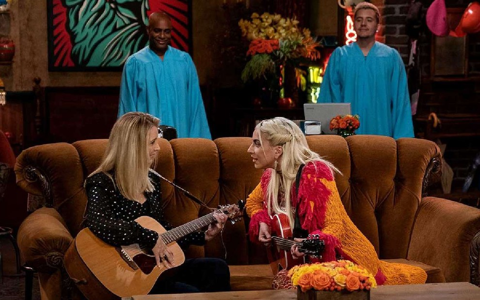 Lisa Kudrow no lado esquerdo e Lady Gaga no lado direito sentadas em sofá e tocando violão com cantores de coral ao redor