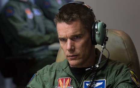 Ethan Hawke com roupas da força aérea, vestindo um grande fone de ouvido com uma feição séria