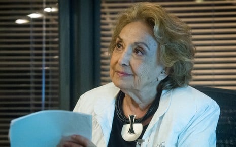 Eva Wilma em traje de médica sentada em uma mesa e segurando papéis com a mão esquerda