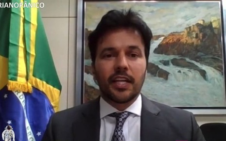 Fábio Faria de terno cinza, camisa e gravata, sentado à frente de uma bandeira do Brasil