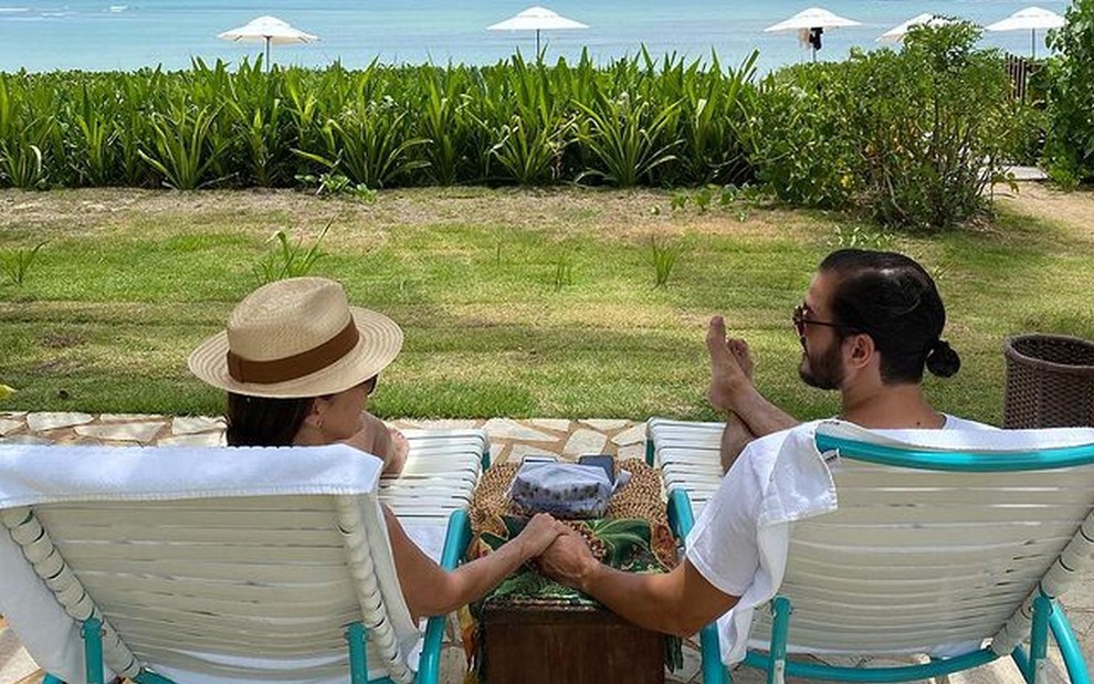 Fátima Bernardes e Túlio Gadêlha de costas, sentados em espreguiçadeiras, à beira da praia