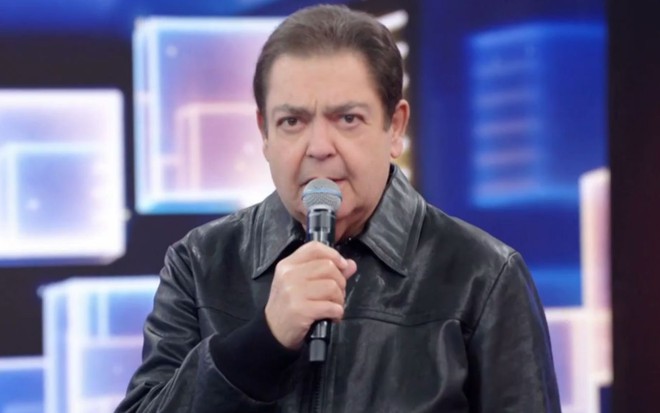 Fausto Silva segura o microfone e está com expressão séria no cenário do Domingão do Faustão (1989-2021)